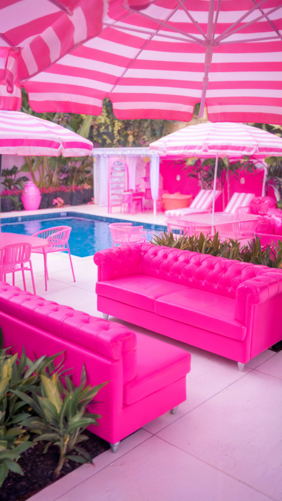 A photo of a Barbie Dreamhouse pool area with a bright pink tufted sofa. There is a pool in the background, and there are pink and white striped umbrellas. There are also pink and white chairs. The floor is made of white tiles. The area is surrounded by lush green plants. The overall ambiance is bright and cheerful.